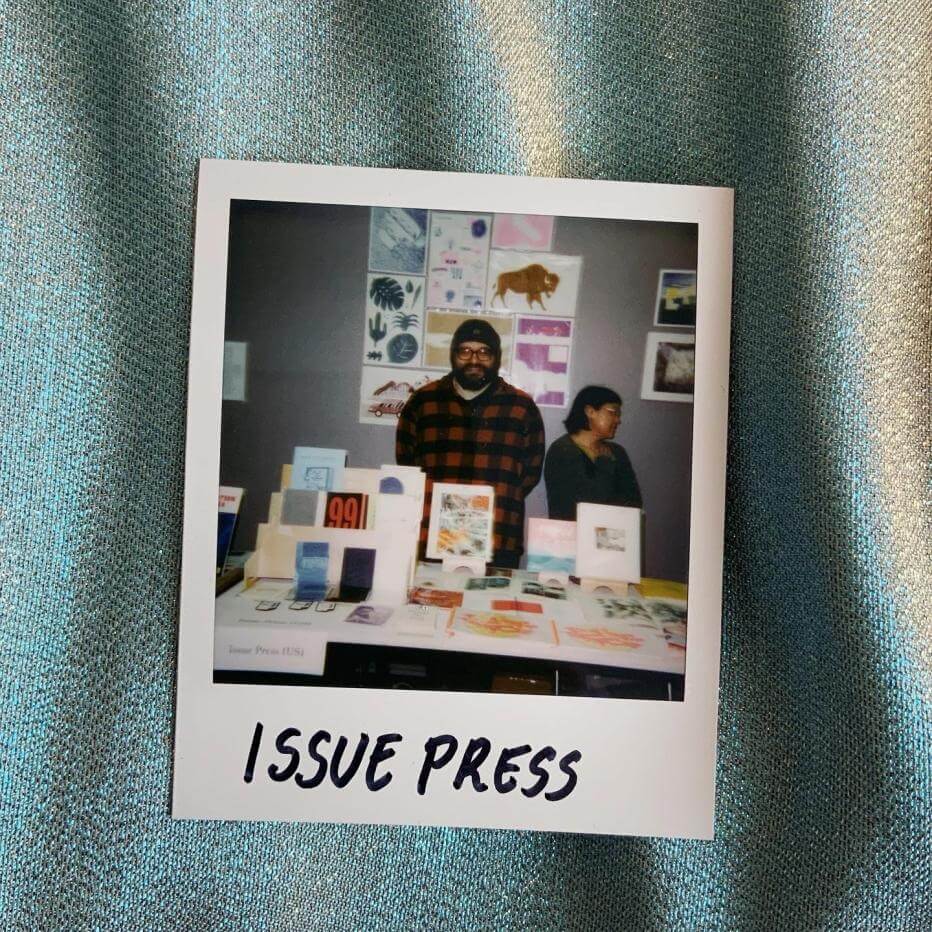 A somewhat meta photo of a polaroid of two people standing between a table full of books and wall covered in prints at an art book fair